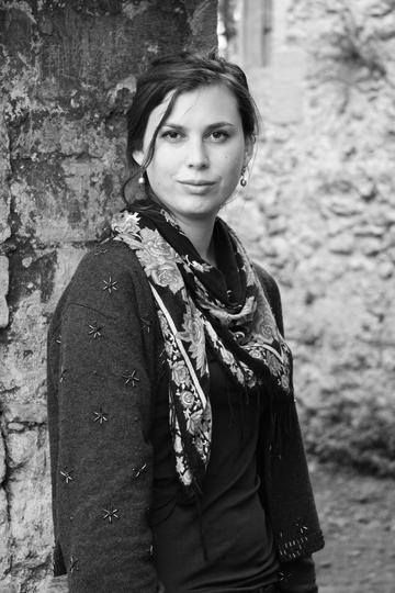 A black & white photograph of Penny Boxall, who stands with her back against a tree trunk. Her face is turned directly towards the viewer, and she is dressed in dark clothing with a bright floral scarf and drop earrings.
