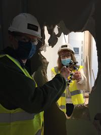 Photo of two people high-vis jackets, helmets and face masks, standing on either side of plasterboard wall and passing a molecular model through a hole in the middle.
